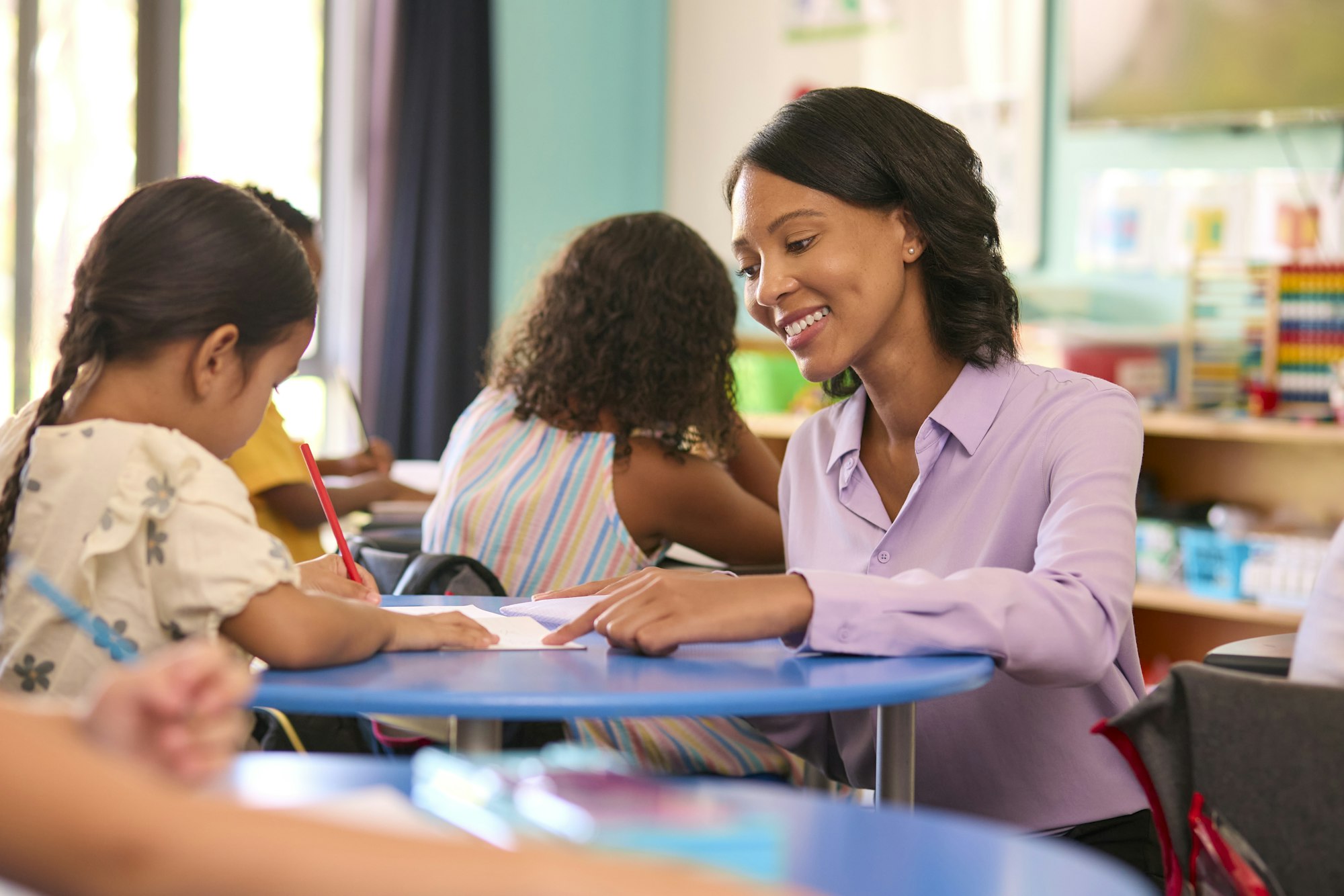 Female Primary Or Elementary School Teacher Helping Students At Desks In Multi-Cultural Class